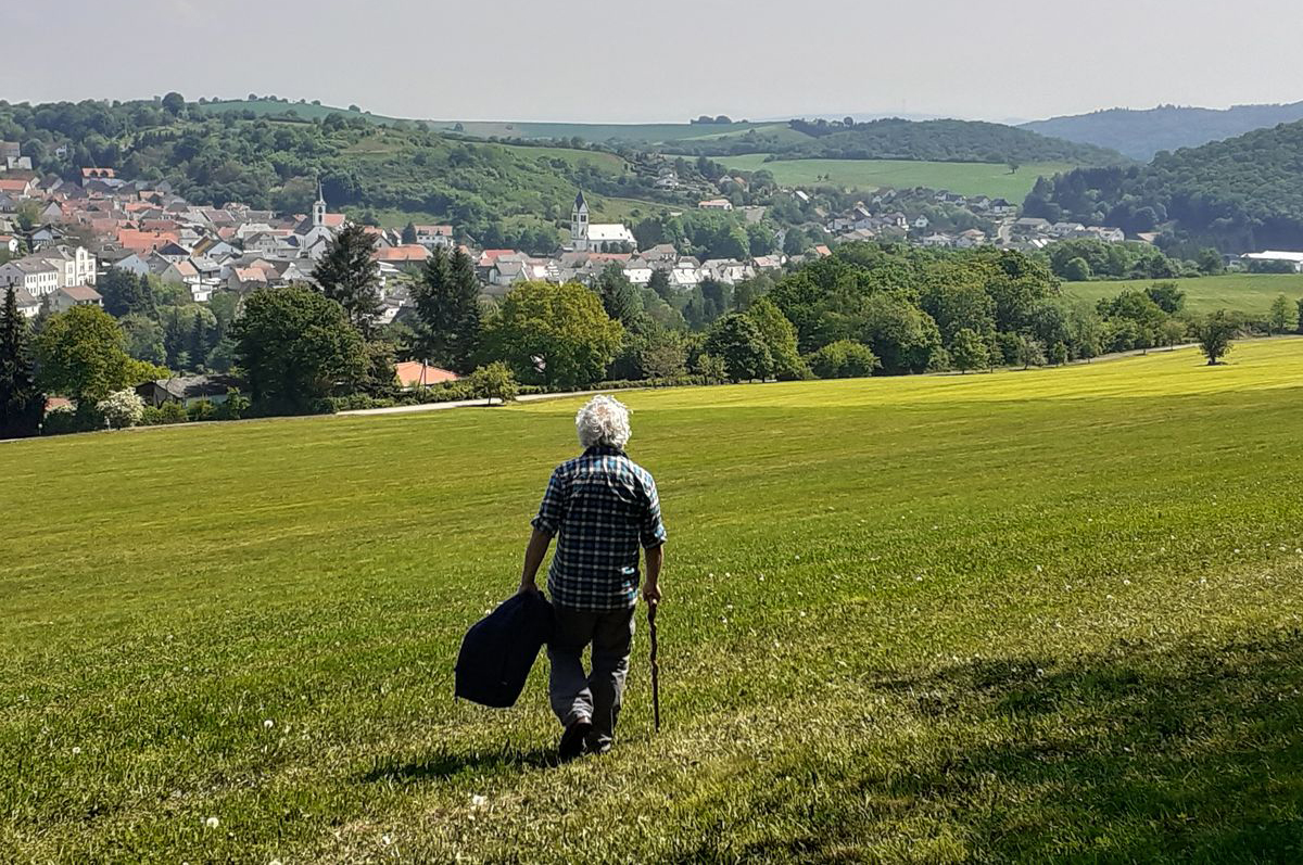 Mann geht mit Stock in der rechten und Jacke in der linken Hand über eine grüne Wiese. In Hintergrund ist ein Dorf zu sehen.