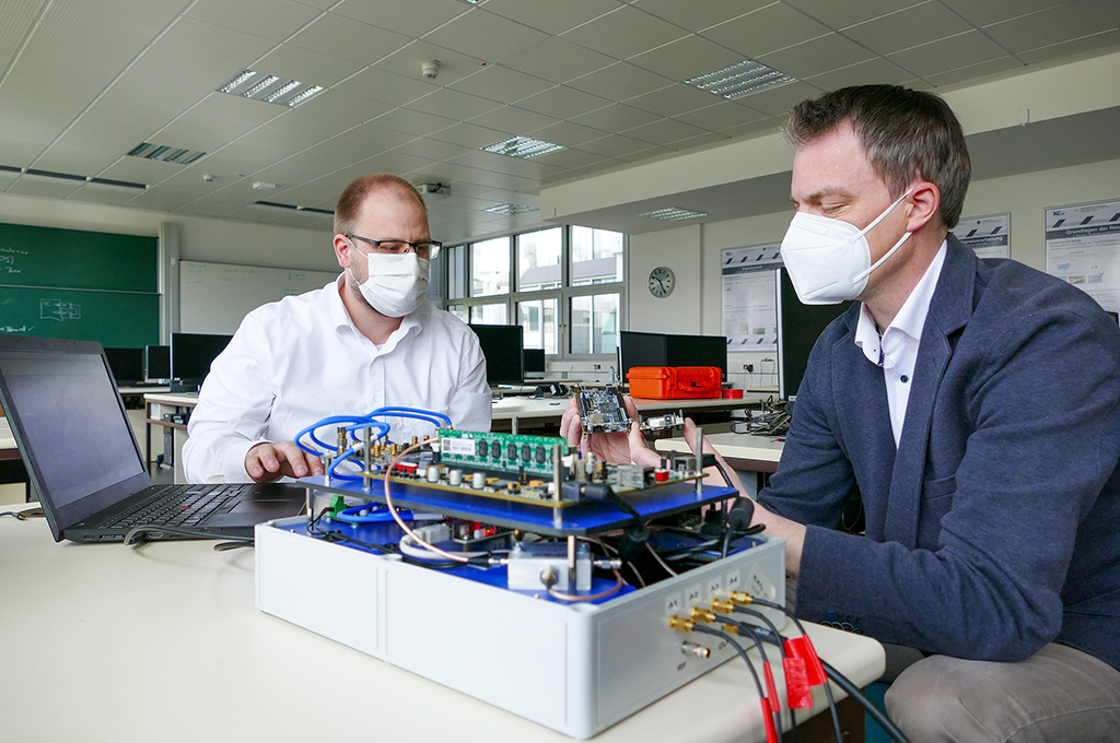 Masterabsolvent Markus Biermann und Professor Kappen sitzen am Tisch und schauen auf ein technisches Gerät, das Biermann gebaut hat.