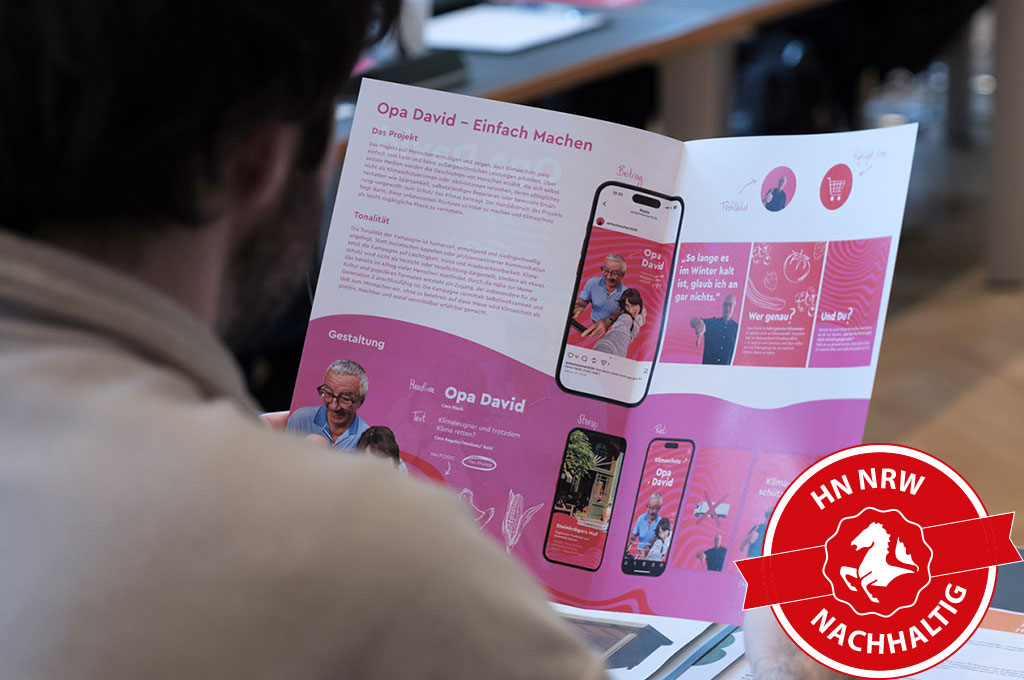 Endpräsentation_Klimaschutz_HENK_86_Slider Eine Person hält eine aufgeschlagene Broschüre mit pinkem Design und dem Titel „Opa David – Einfach machen“, auf der Texte, Fotos und Smartphone-Darstellungen zu sehen sind.