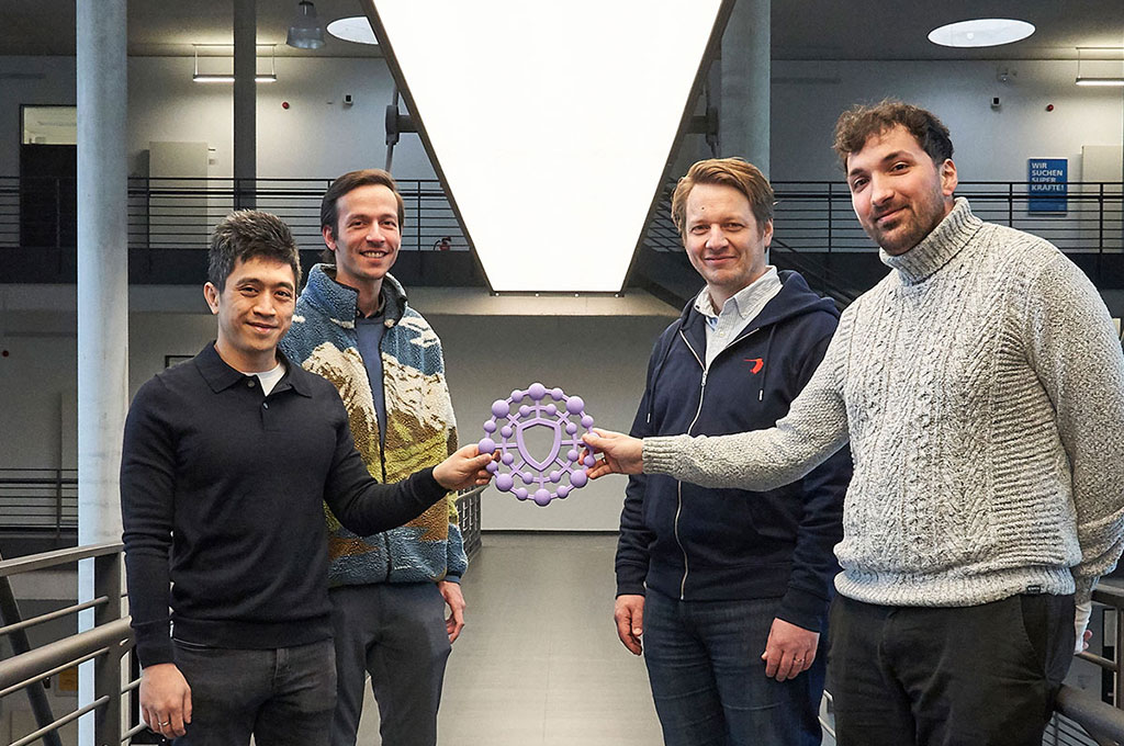 Vier erwachsene Personen stehen in einem modernen, hellen Innenraum mit Betonstützen und einer Galerie. Zwei Personen halten gemeinsam ein rundes, lilafarbenes Objekt mit einer schildähnlichen Form in der Mitte. Alle blicken in die Kamera und wirken freundlich. Im Hintergrund sind Geländer, Deckenleuchten und offene Ebenen des Gebäudes zu sehen.